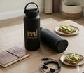 Black water bottles with 'mi MINDFUL' branding on a wooden table with plates of food and a notebook.