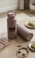 Pink water bottle with 'mi MINDFUL' branding on a wooden surface with plates of food and earphones.