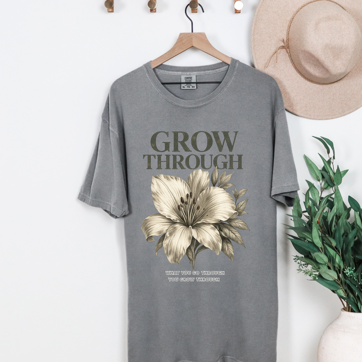 Gray t-shirt with floral design and text on a hanger with a hat and plant in the background