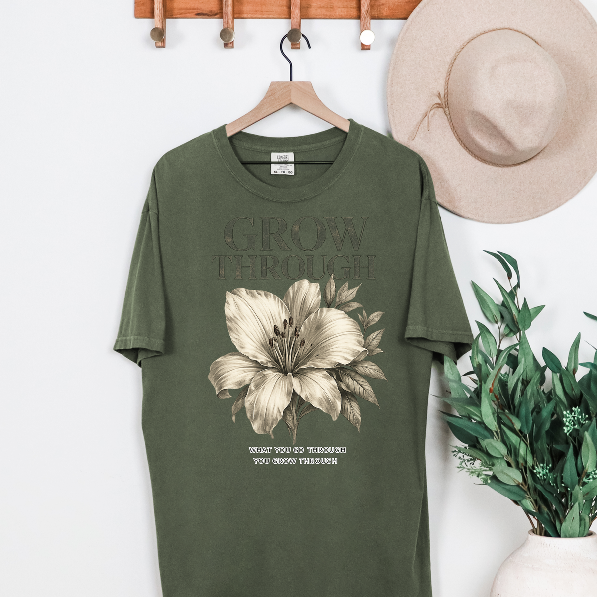 Green t-shirt with floral design and text on a hanger, next to a hat and plant.